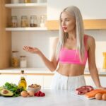 Gorgeous platinum blonde woman in a neon pink crop top with bare midriff standing at a bright marble kitchen counter comparing a keto food spread with avocado nuts and olive oil against a carnivore food spread with ribeye steak salmon and butter representing the keto vs carnivore diet choice for weight loss
