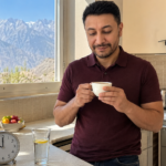Shahbaz Ahmed founder of KetoFramework in a dark teal crew neck sitting outdoors at his Gilgit-Baltistan afternoon table with a hand-drawn 18:6 intermittent fasting schedule notepad showing fasting 18 hours and eating 6 hours with a cup of black tea looking directly at camera with a warm confident smile representing 18:6 intermittent fasting and how it differs from 16:8
