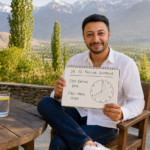 Shahbaz Ahmed founder of KetoFramework in a white shirt sitting outdoors at his Gilgit-Baltistan morning table holding a handwritten 14:10 fasting schedule notepad showing stop eating 8pm and first meal 10am looking directly at camera with a warm encouraging smile representing 14:10 intermittent fasting and why it is the gentlest starting protocol for IF beginners